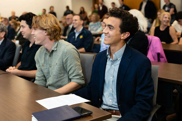 young man in suit smiling during lecture with other people in the background