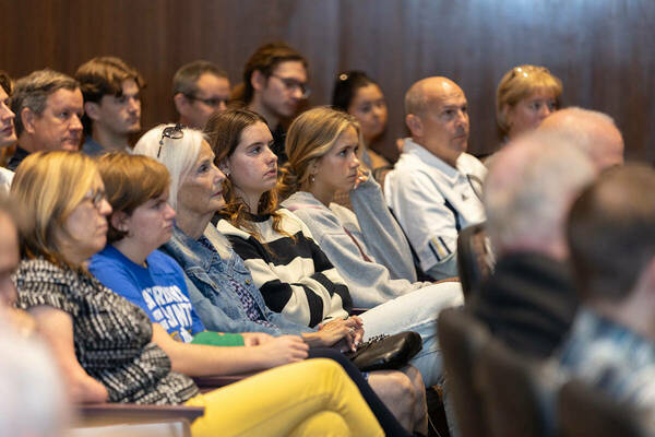 An audience attentively watches a presentation in a lecture hall with wood-paneled walls. Individuals are seated in rows, some wearing Notre Dame apparel.