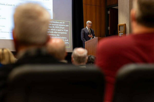 Dr. Jay Bhattacharya stands at a podium, gesturing with his hand as he addresses an audience. A projected presentation is visible behind him, partially obscured by the heads of audience members in the foreground.
