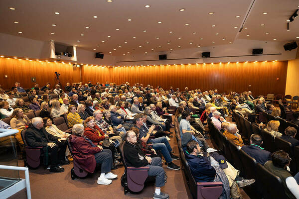 A large audience sits in a lecture hall with wood-paneled walls and burgundy seats. The audience members, dressed in a variety of attire, are attentively watching a presentation.
