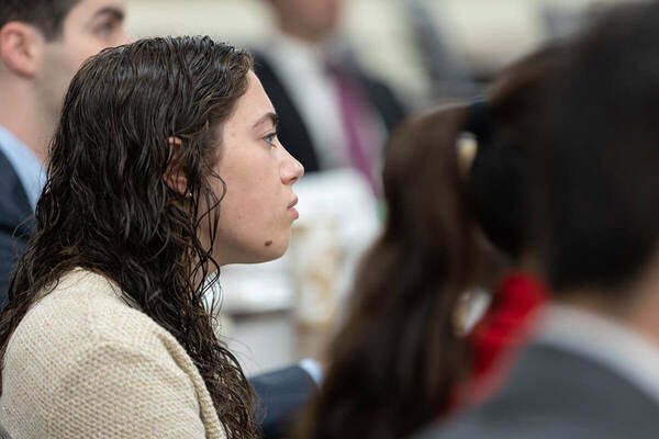 A student with long, dark, wavy hair listens attentively in profile view. She wears a cream-colored sweater. Other attendees are slightly blurred in the background.