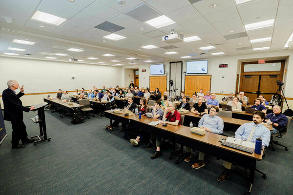 Josiah Ober in a dark suit gestures while addressing an audience seated at tables in a lecture hall.  Two screens display presentation slides, and video cameras record the event.