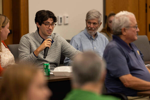 A man in a gray sweater speaks into a microphone during a meeting or presentation.  Other attendees listen.