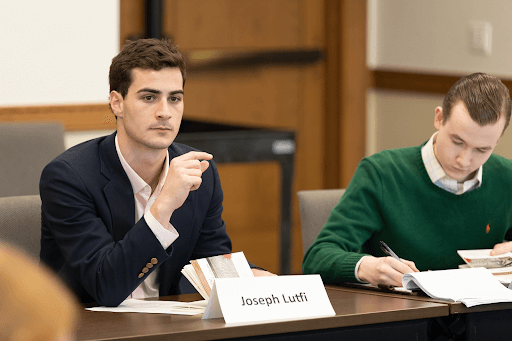 Joseph Lutfi gestures while speaking in a classroom setting. Another student in a green sweater writes notes beside him.