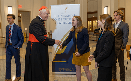 A cardinal, wearing a red sash and black cassock, shakes hands with a woman in a gold dress and black blazer. Other students in business attire stand nearby in a University of Notre Dame building lobby. A banner for the Center for Citizenship & Constitutional Government stands between the cardinal and the students.