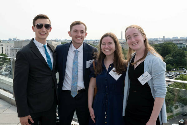Four Notre Dame students wearing professional attire smile on a rooftop overlooking the Washington, D.C. skyline, including the Washington Monument.