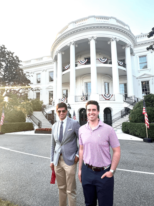 Two men stand in front of the White House, decorated with American flags.  One wears a gray sport coat and khaki pants, the other a pink gingham polo shirt and dark pants. They are smiling.