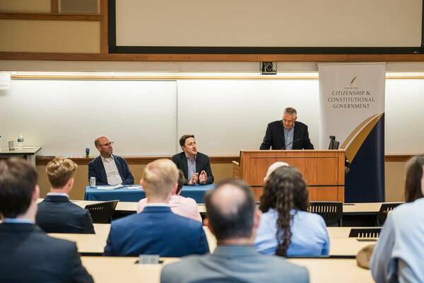 A panel discussion features three men. One with gray hair speaks from a wooden podium. Another, in a blue blazer, gestures from a table while a third, bald man in a checkered shirt, listens. A banner for the Center for Citizenship & Constitutional Government is visible.