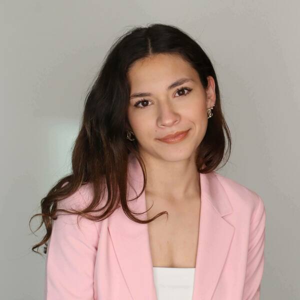 A young woman with long dark brown hair smiles gently, wearing a pink blazer and small silver interlocking C earrings.