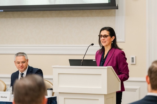 A smiling woman in a magenta blazer and glasses speaks at a white podium with a microphone. A man with grey hair in a dark suit sits nearby, looking down.