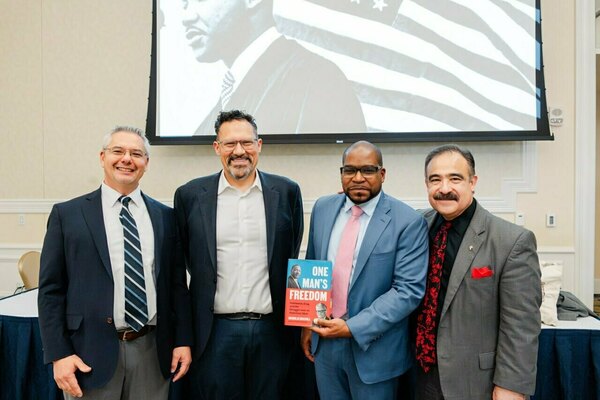 Four smiling men in suits stand before a screen displaying a black and white image of Martin Luther King Jr. A Black man in a light blue suit and pink tie holds a book titled 'One Man's Freedom.' Another wears a grey suit, black shirt, and red tie.