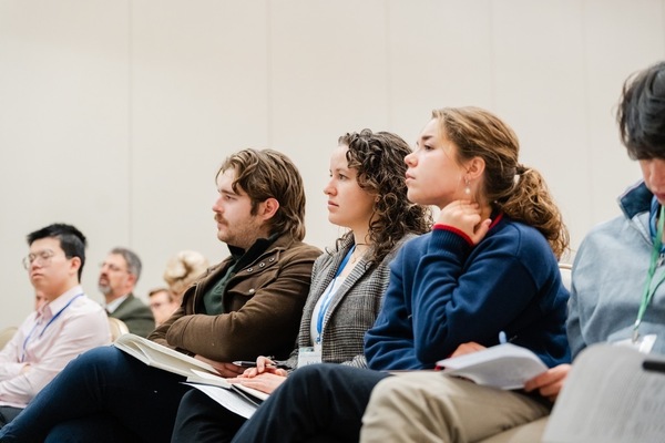 Three people sit attentively in a conference room. A man in a brown jacket holds a notebook, a woman in a plaid blazer holds a pen, and another woman in a dark blue sweatshirt with red trim touches her neck, all looking focused.