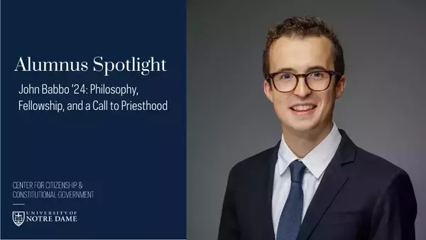 John Babbo '24, a young man with curly brown hair and glasses, smiles brightly, wearing a dark suit, white shirt, and blue tie against a grey background.