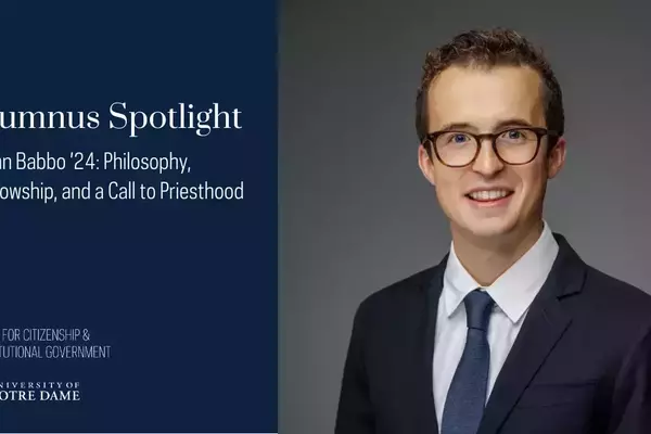 John Babbo '24, a young man with curly brown hair and glasses, smiles brightly, wearing a dark suit, white shirt, and blue tie against a grey background.