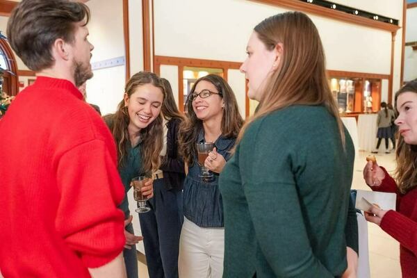 A man in a red sweater talks to two smiling women at an indoor event. A woman with long curly hair laughs, holding a drink. Beside her, a woman in glasses smiles, also holding a drink. Another woman in dark green listens, and a woman in red eats a pastry.