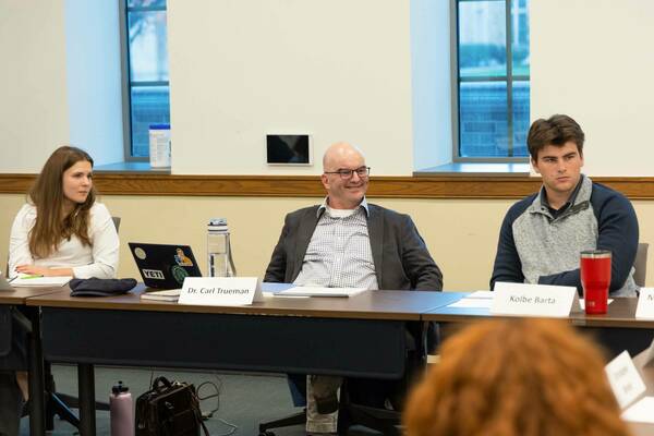 Dr. Carl Trueman, smiling, sits at a conference table between a woman in a white shirt and Kolbe Barta in a navy and grey sweater. All look right.
