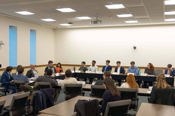 About 20 students and one older man with glasses sit around a U-shaped conference table in a well-lit room, participating in a discussion. Name placards are visible.