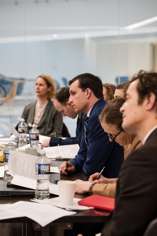People sit around a conference table with papers and water bottles. A man in a blue suit and tie holds a pen, looking forward attentively, as others around him take notes.