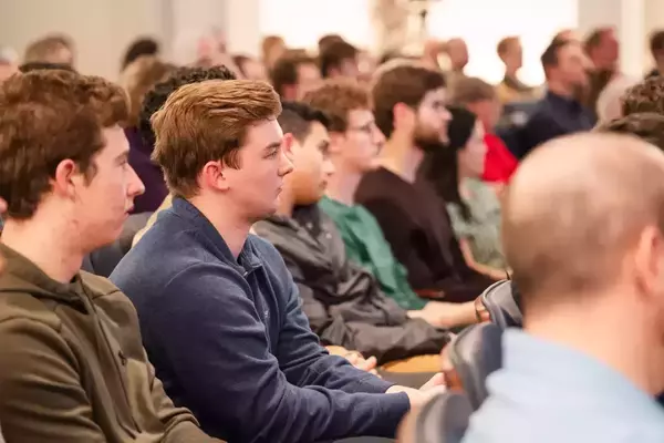 Students in a lecture hall. A young man in a blue sweater and another in a green hoodie watch attentively.