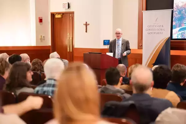 Man in grey suit and blue tie smiles, speaks at podium to audience. A crucifix and Center for Citizenship banner are behind him.