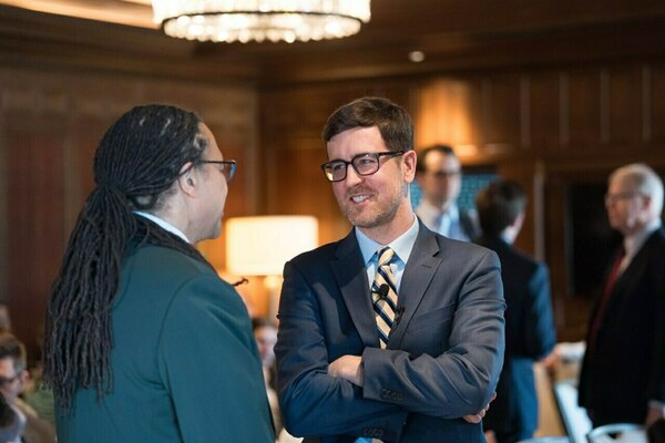 Man with dreadlocks speaks to a smiling man in a blue suit with a striped tie and crossed arms at an indoor event.