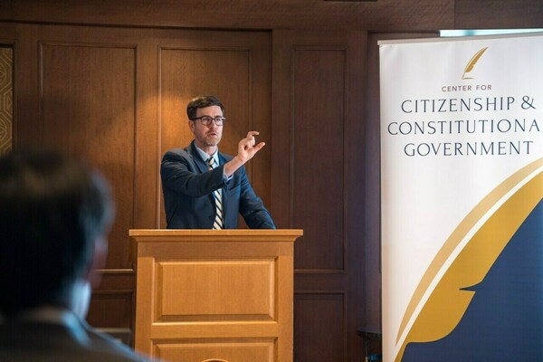 Man in blue suit, striped tie, speaks and gestures at a wooden podium. A Center for Citizenship & Constitutional Government banner is visible.