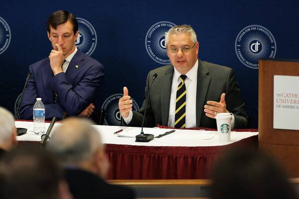 Two men at a Center for the Constitution event. One in gray suit and striped tie speaks, gesturing. The other in blue suit looks thoughtful.