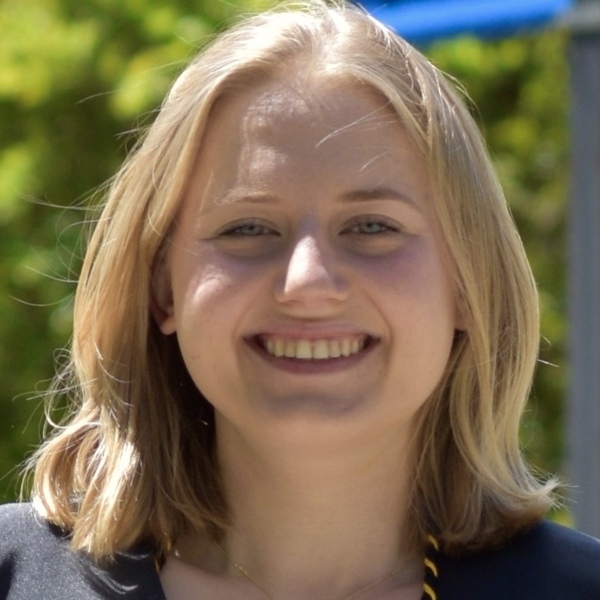 Smiling young woman with blonde hair and blue eyes, wearing a dark graduation gown with a gold honor cord.