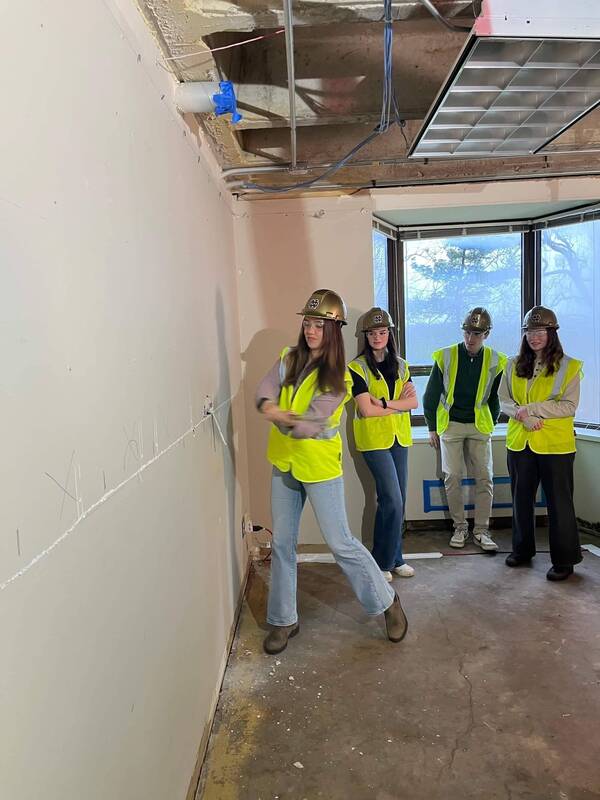 Four people in gold Notre Dame hard hats and neon vests stand in a room under renovation. One gestures, others observe.